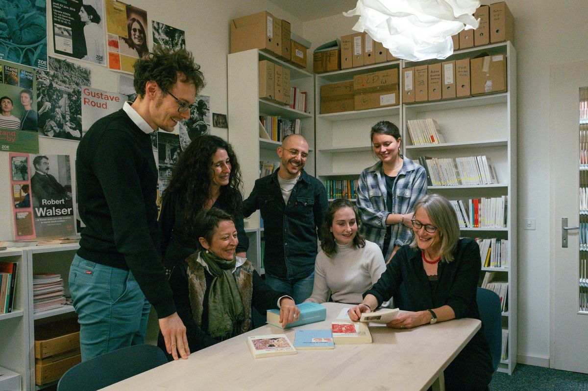 Groupe de personnes autour d’une table dans une bibliothèque, regardant des livres sur la table. Des étagères remplies de livres et de boîtes d’archives sont visibles en arrière-plan.