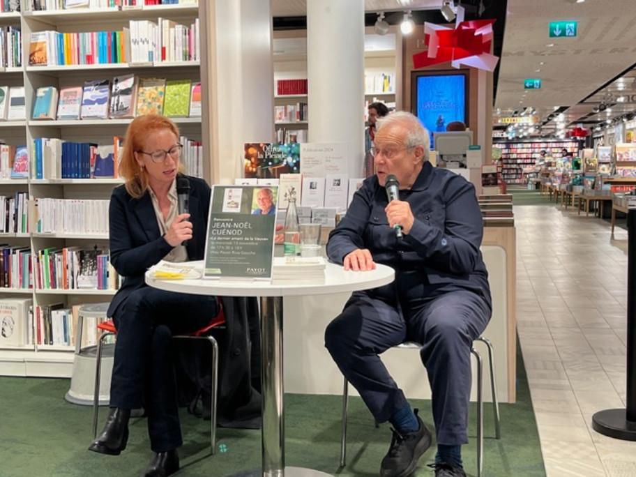 Deux personnes discutent lors d’une séance de dédicace dans une librairie, avec des livres en arrière-plan.