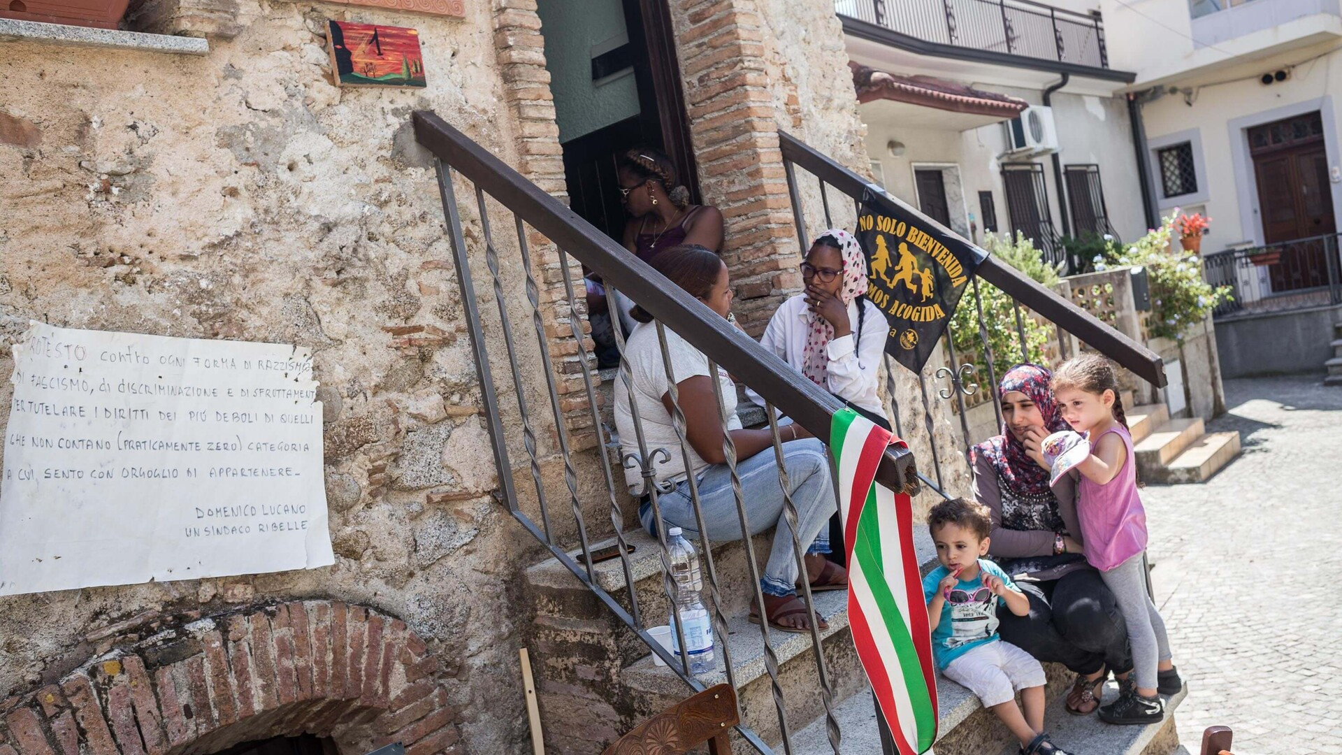 Un groupe de personnes assis sur les marches d’une maison en pierre, discutant et souriant. Un enfant joue avec une poussette tandis qu’un drapeau italien est accroché à la rambarde. Une pancarte est fixée au mur avec un texte en italien.