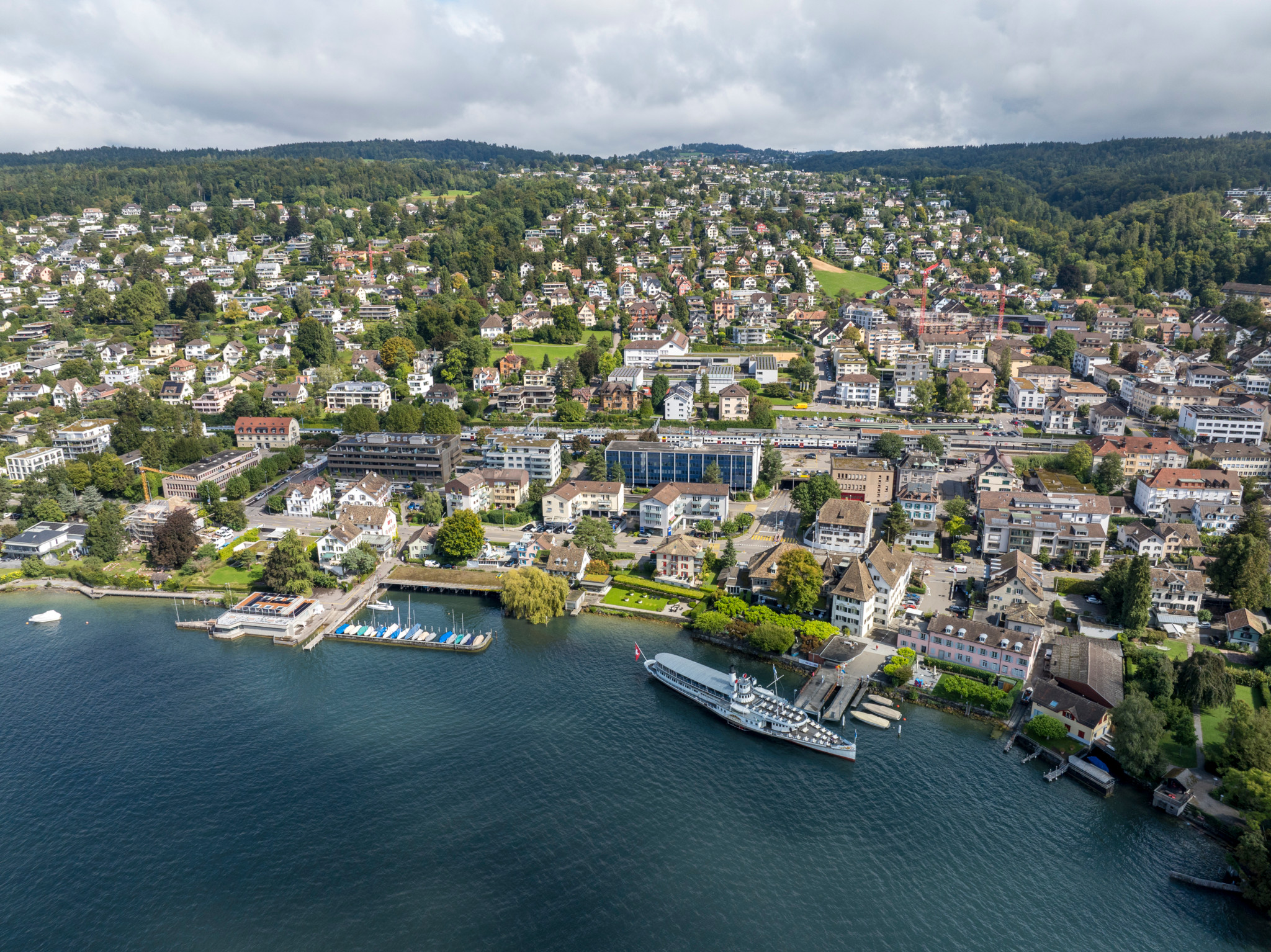 Luftaufnahme von Küsnacht am Zürichsee mit Seepromenade, angelegtem Dampfschiff, Hafen und bewaldeten Hügeln im Hintergrund.
