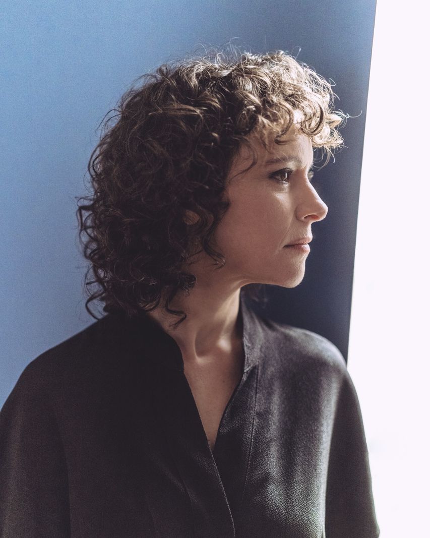 Une femme avec des cheveux bouclés, portant une chemise foncée, regarde par une fenêtre lumineuse.