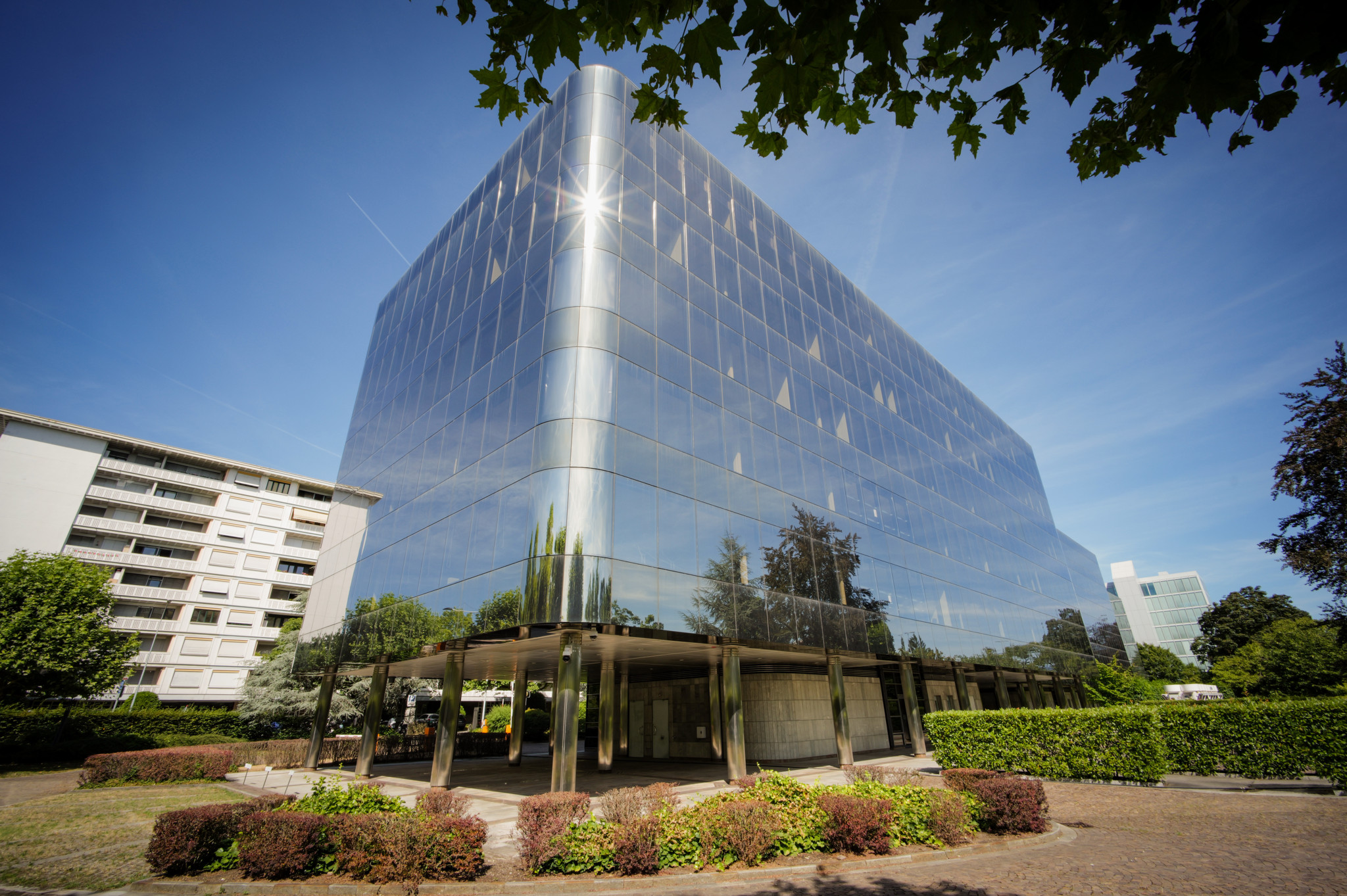 Immeuble de bureaux à façade vitrée réfléchissante au 2-6 Avenue des Morgines, Esserts, Genève, entouré de végétation sous un ciel bleu. Immeuble de bureaux à façade vitrée réfléchissante au 2-6 Avenue des Morgines, Esserts, Genève, entouré de végétation sous un ciel bleu.