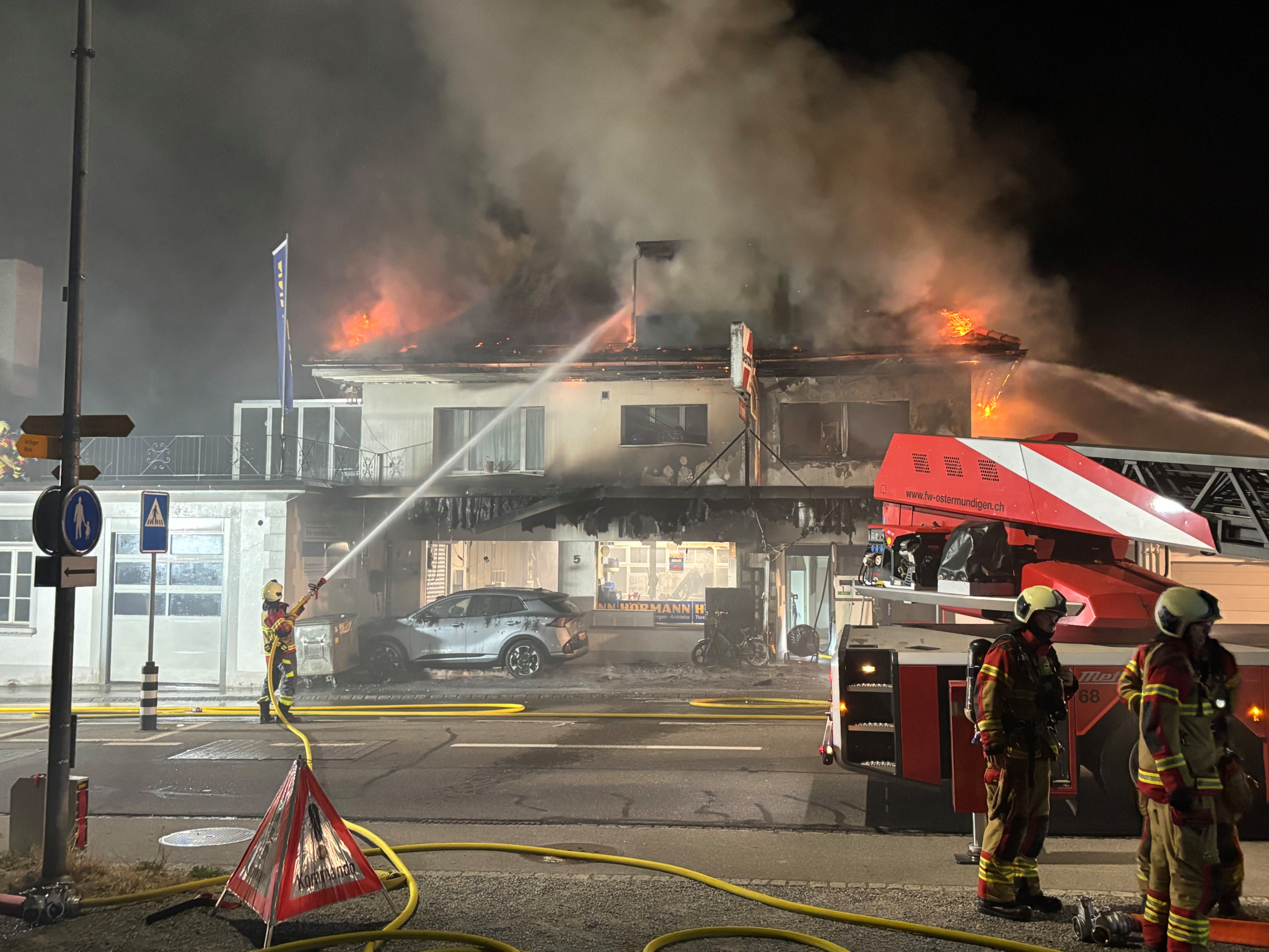 Feuerwehr mit Drehleiter vor einem ausgebrannten Gebäude mit zerstörtem Dach neben einer Benzin-Diesel-Tankstelle.