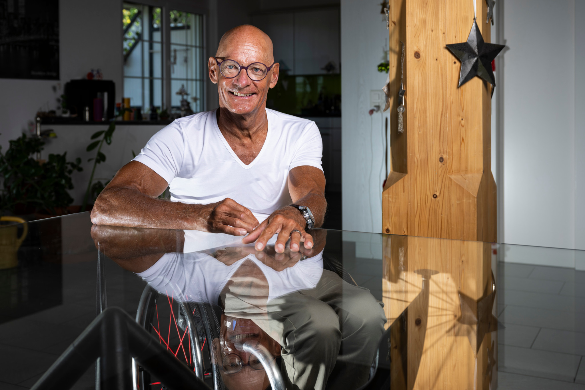 Älterer Mann im Rollstuhl sitzt an einem Glastisch in einem modernen Wohnraum mit Pflanzen im Hintergrund.
