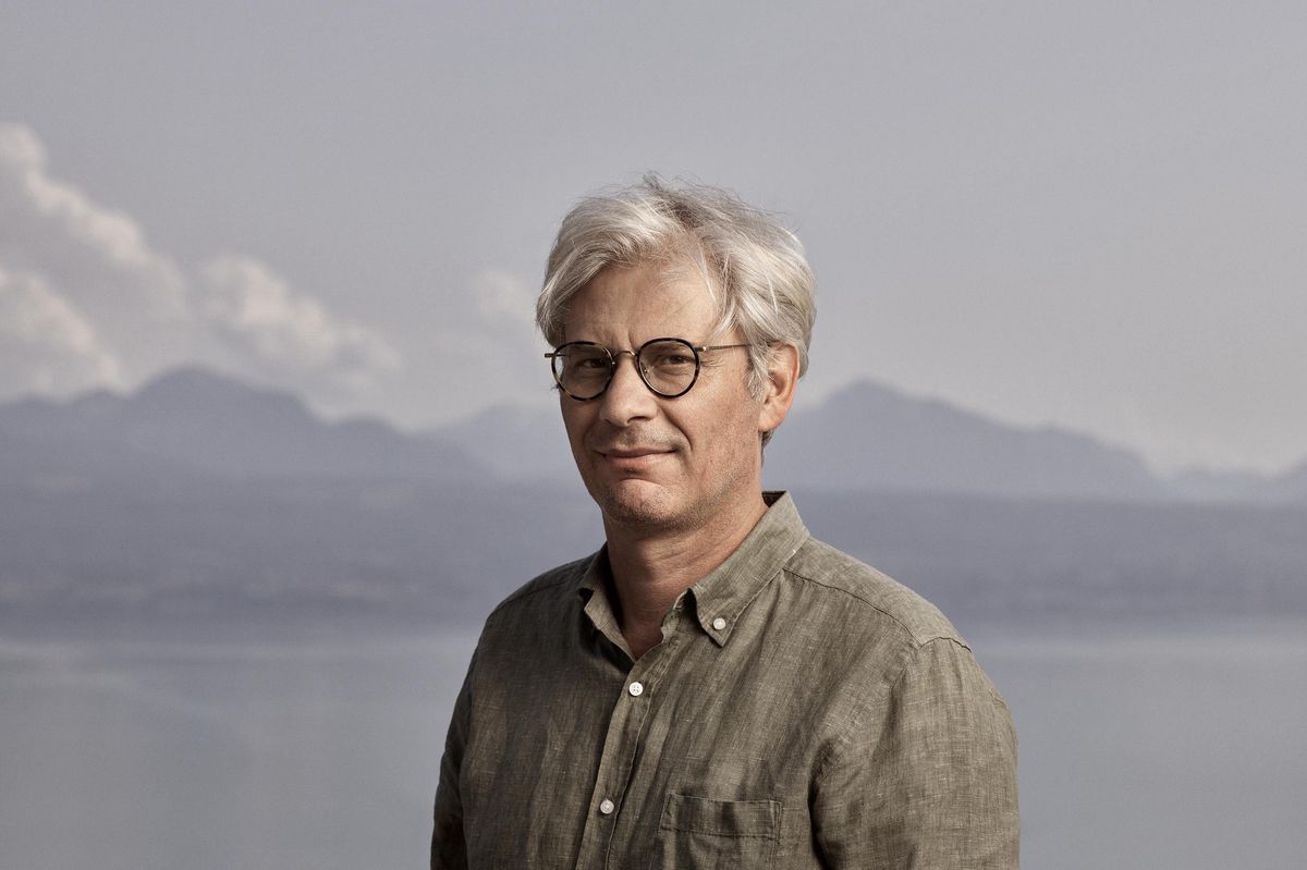 Homme aux cheveux gris portant des lunettes et une chemise verte, debout devant un lac avec des montagnes en arrière-plan.