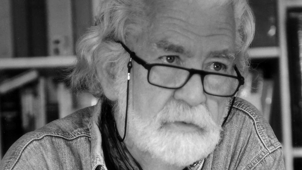 Homme âgé avec des lunettes, cheveux blancs et barbe, portant une chemise en jean, en noir et blanc.