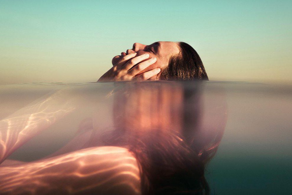 Personne se reposant à moitié immergée dans l’eau, visage tourné vers le ciel avec une lumière douce et diffusée.