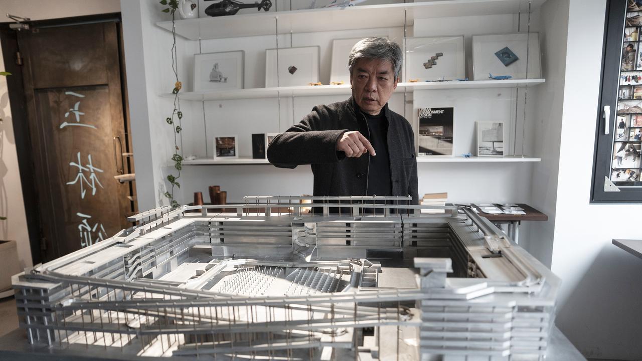 Un homme examine une maquette architecturale complexe sur une table dans une pièce remplie de documents et de dessins techniques.