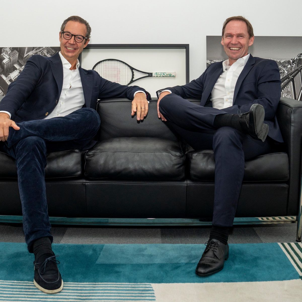 Deux hommes en costumes assis sur un canapé en cuir noir, avec des œuvres d’art en arrière-plan.