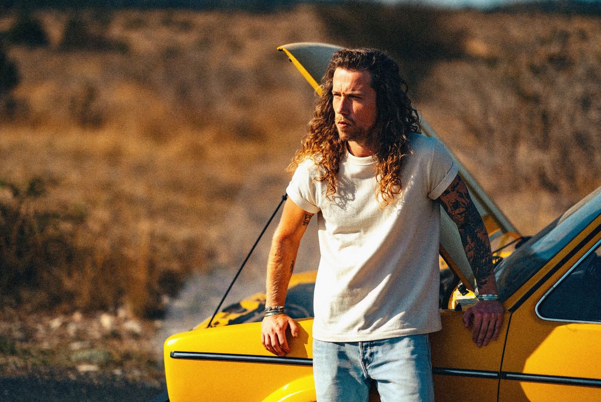 Homme aux cheveux bouclés debout près d’une voiture jaune avec le capot ouvert sur une route isolée.