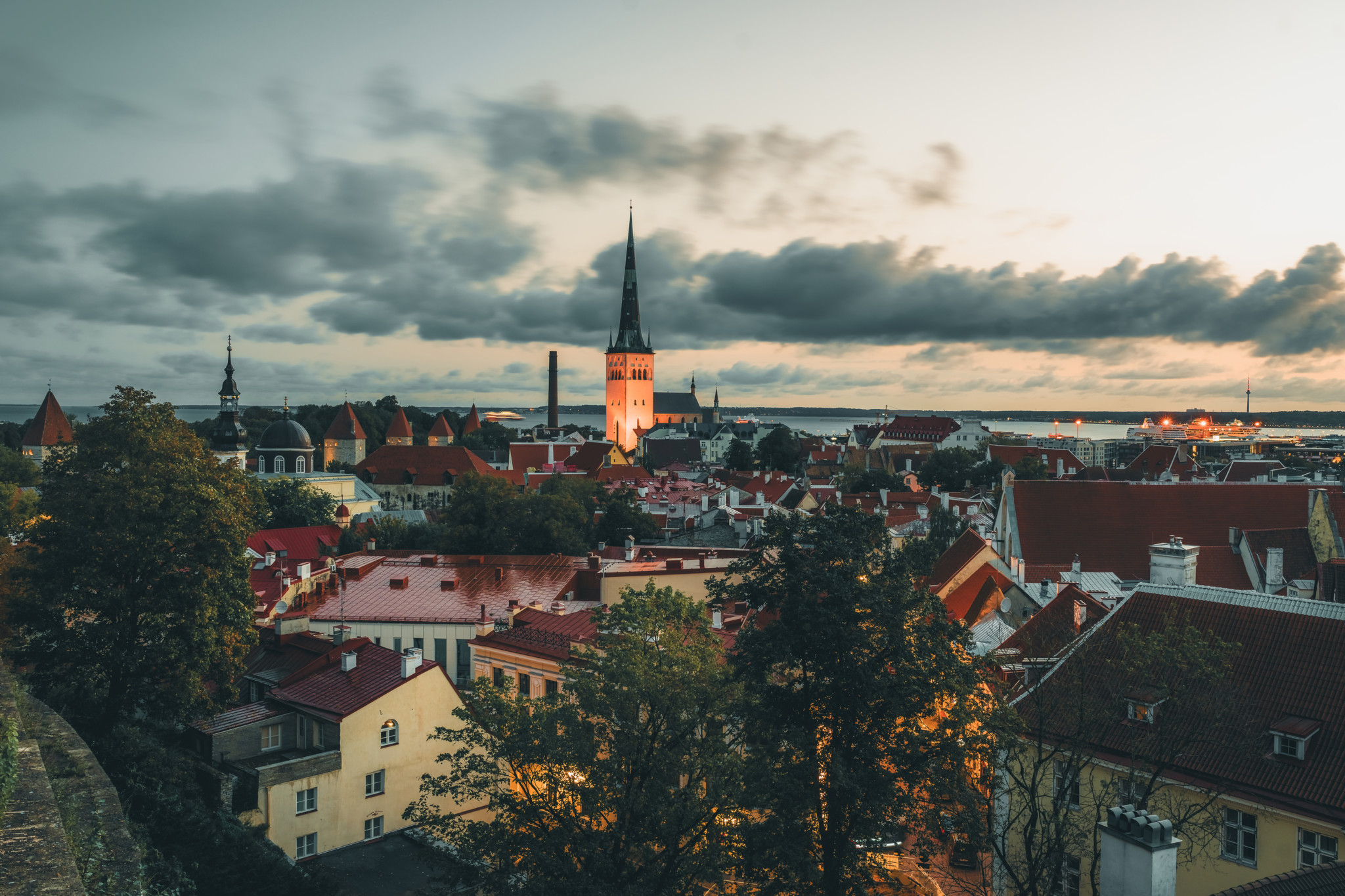Panoramablick über die Altstadt von Tallinn mit dem beleuchteten Turm der Olaikirche unter bewölktem Abendhimmel.