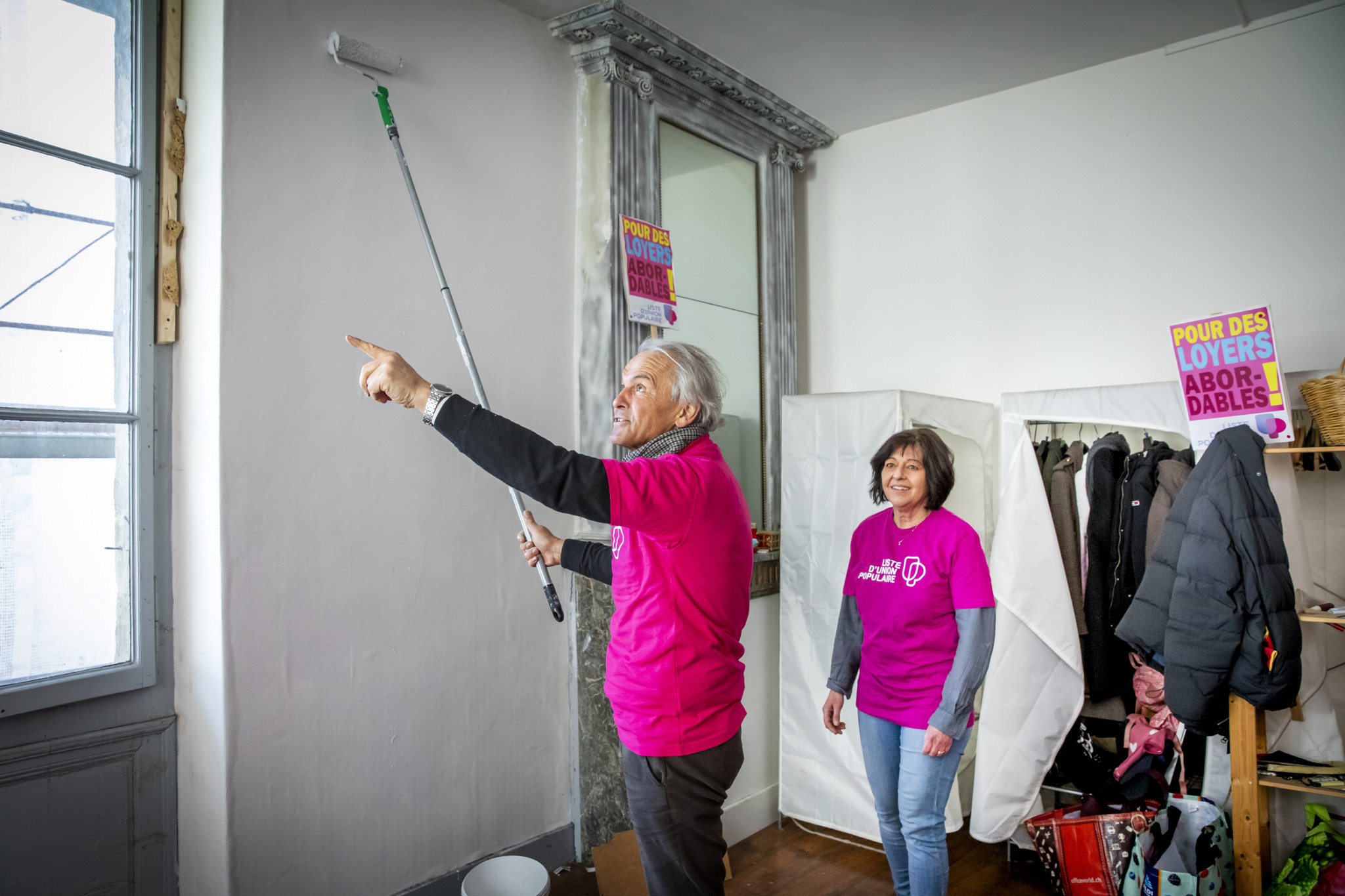 Un homme et une femme peignent un mur blanc avec des rouleaux, portant des t-shirts roses. Des affiches pour des loyers abordables sont visibles.
