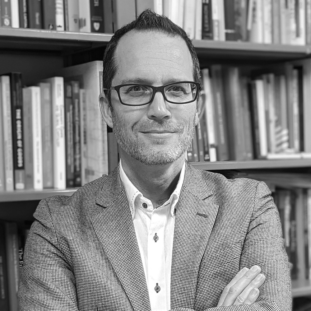 Homme souriant portant des lunettes et une veste à carreaux, debout devant une étagère remplie de livres.