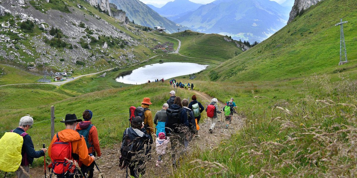Groupe de randonneurs descendant un sentier de montagne à Leysin, avec un petit lac et des montagnes en arrière-plan.