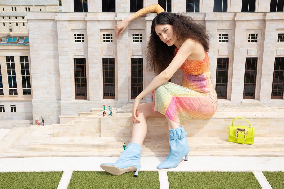 Femme aux cheveux bouclés posant devant un bâtiment en tenue colorée avec des bottes bleues et un sac jaune vif.