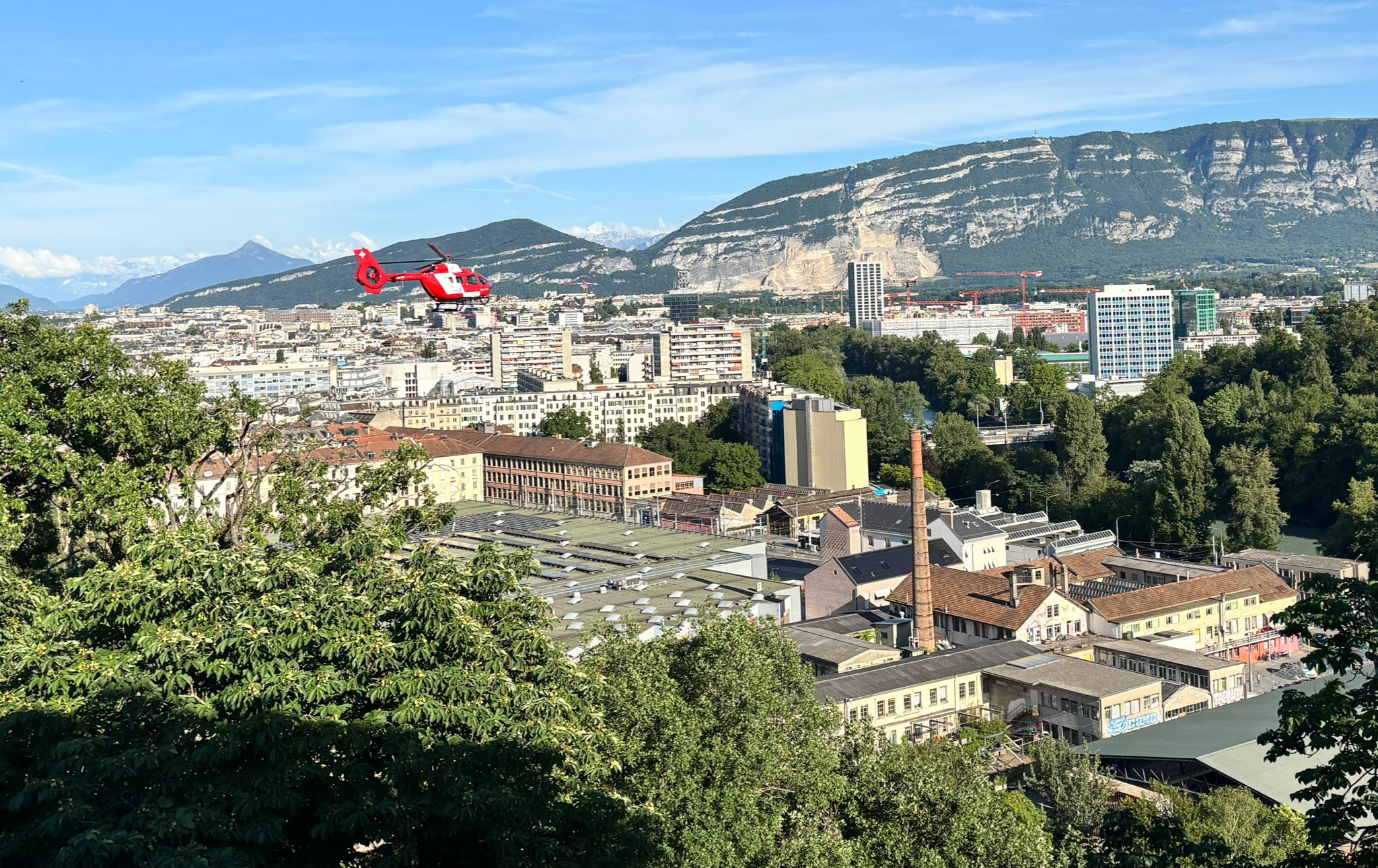 Un hélicoptère rouge survole le paysage urbain de Genève, avec les Alpes et le Salève en arrière-plan par temps ensoleillé. Un hélicoptère rouge survole le paysage urbain de Genève, avec les Alpes et le Salève en arrière-plan par temps ensoleillé.