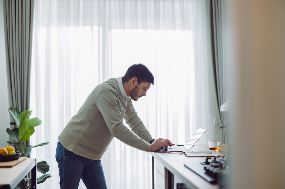 Homme debout dans une cuisine, utilisant un ordinateur portable sur un comptoir, avec une plante verte et des rideaux blancs en arrière-plan.