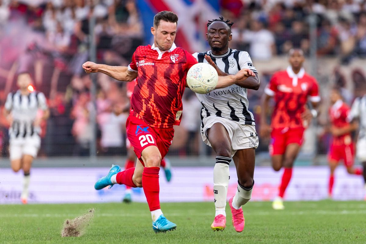 Deux joueurs de football en action lors d'un match, l'un portant un maillot rouge, l'autre un maillot blanc et noir, disputant le ballon sur le terrain.