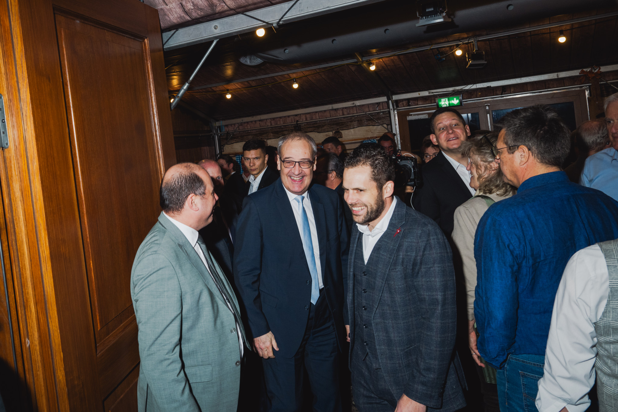 Eine Gruppe von lächelnden Männern in Anzügen steht in einem belebten Raum, einer trägt eine Krawatte.