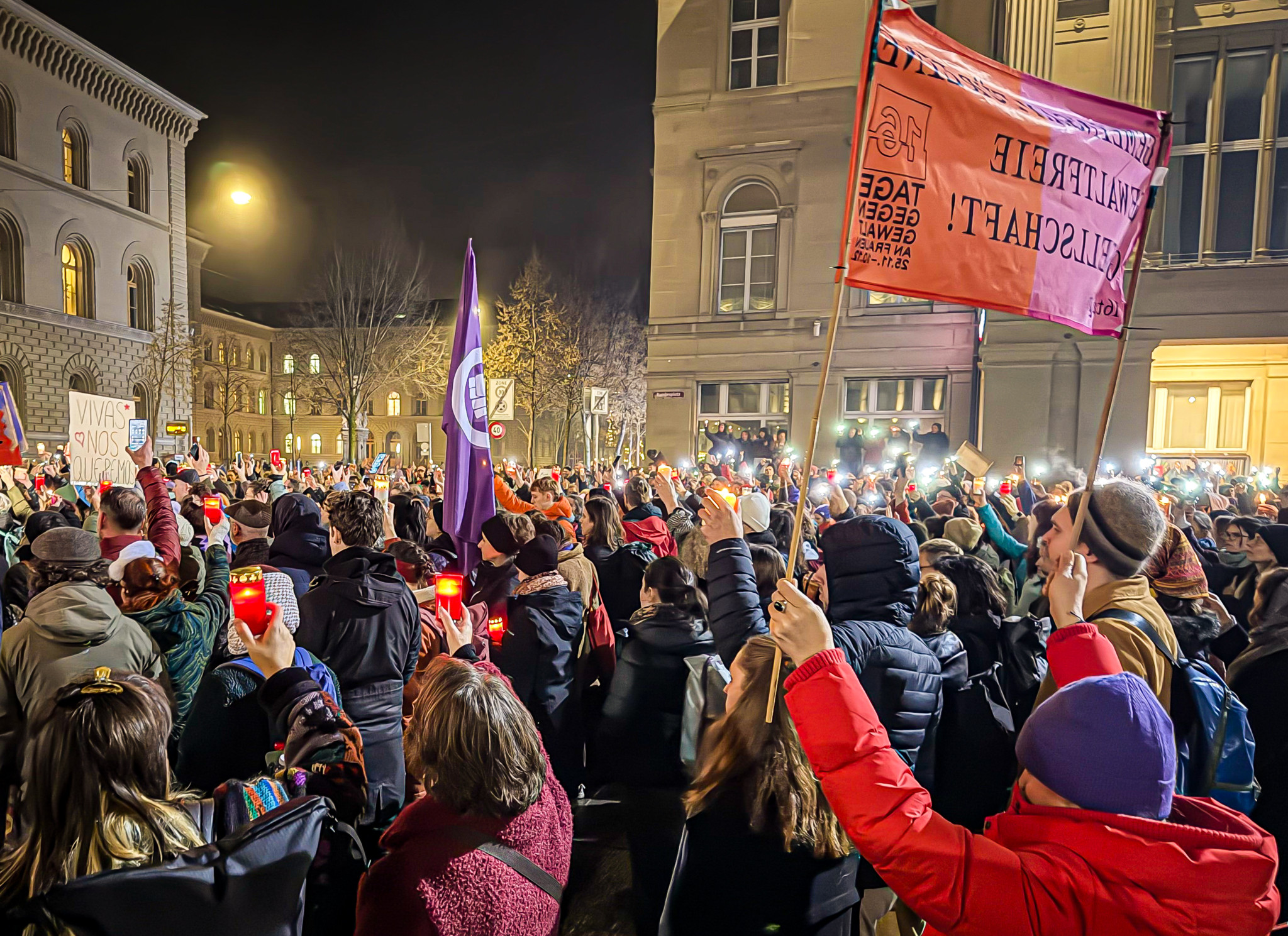 Hunderte von Menschen protestieren bei Nacht auf dem Bundesplatz in Bern, mit Fahnen und Lichtern.