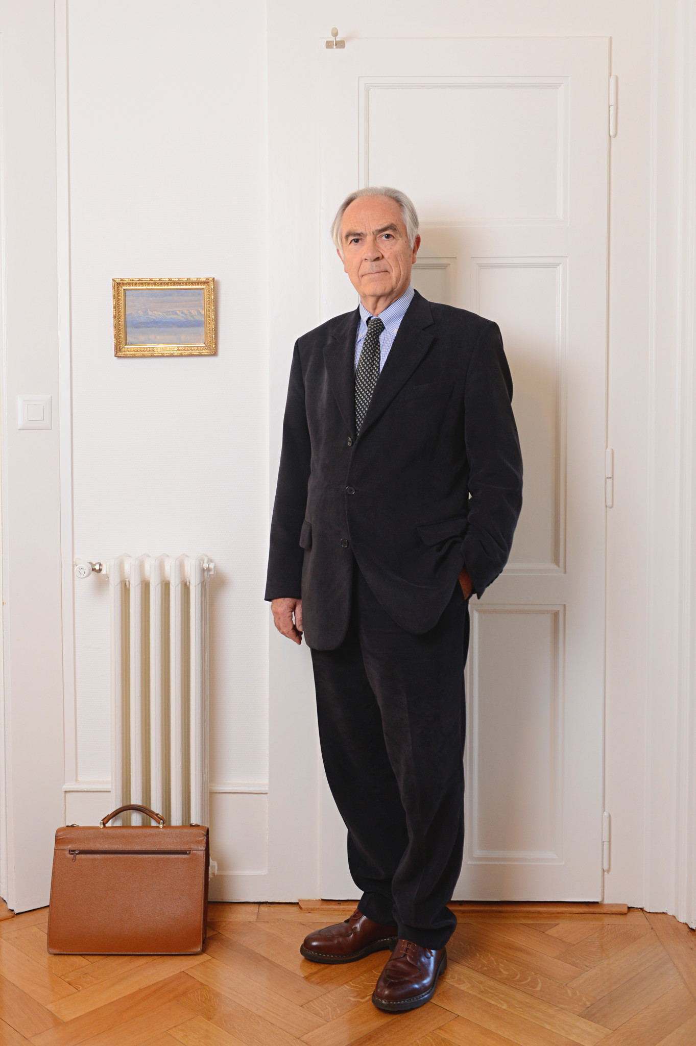Christian Nils Robert debout dans une pièce, vêtu d’un costume sombre, avec une valise en cuir posée au sol, Genève, 29 octobre 2012.