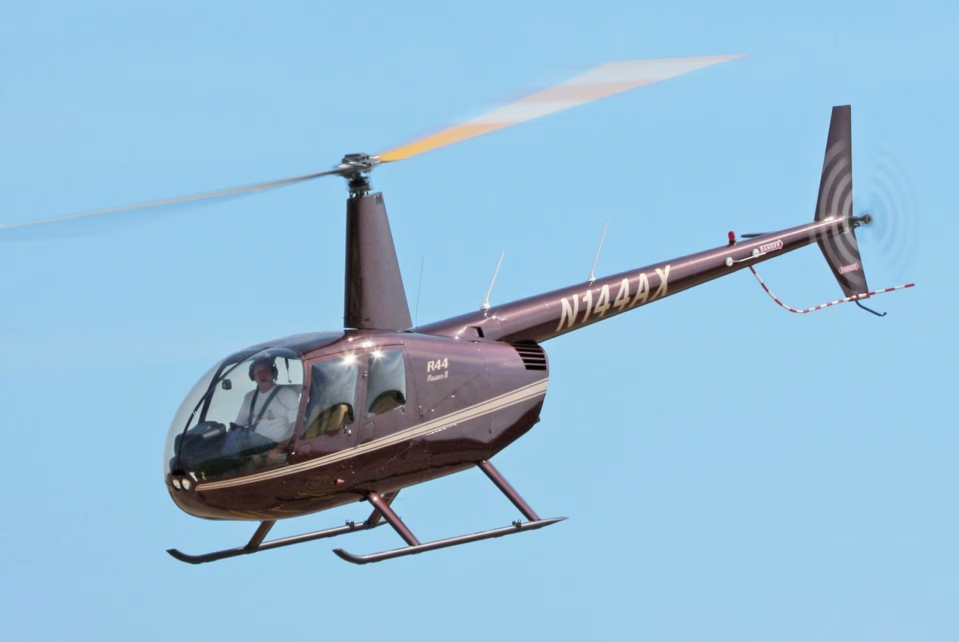 Ein brauner Hubschrauber vom Typ Robinson R44 mit der Kennung N744AX fliegt gegen einen klaren blauen Himmel.