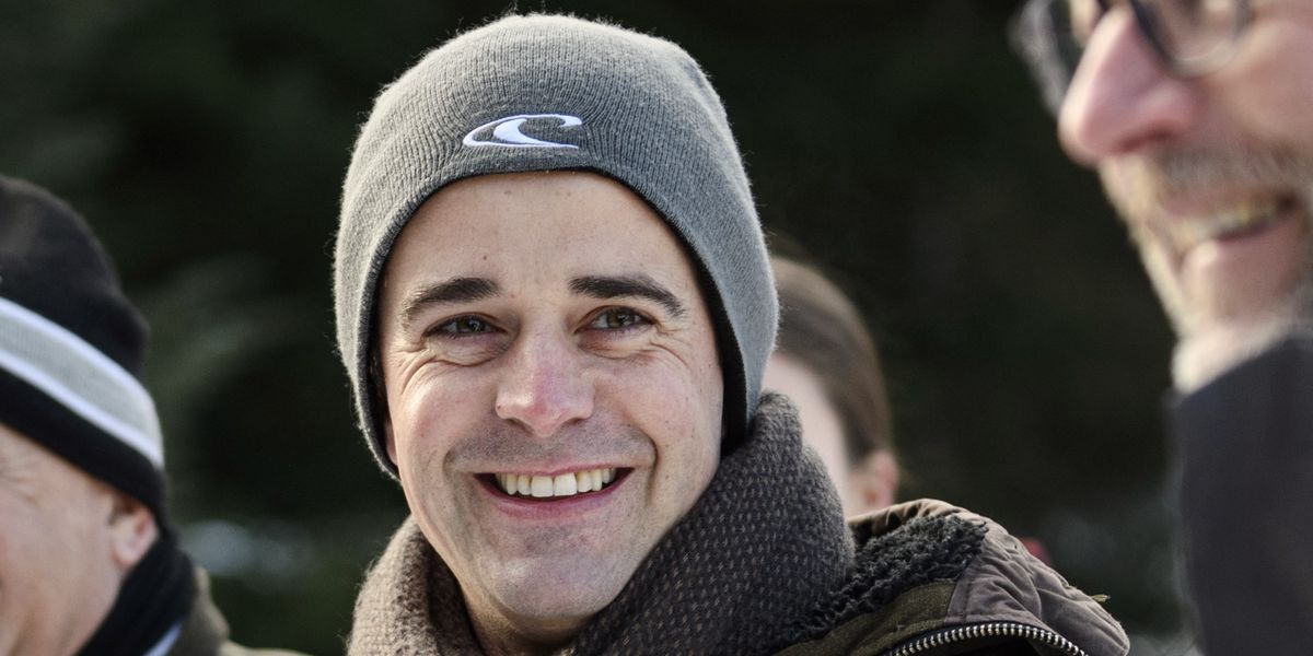 Mann mit grauer Mütze lächelt im Freien, während er von anderen Personen umgeben ist.