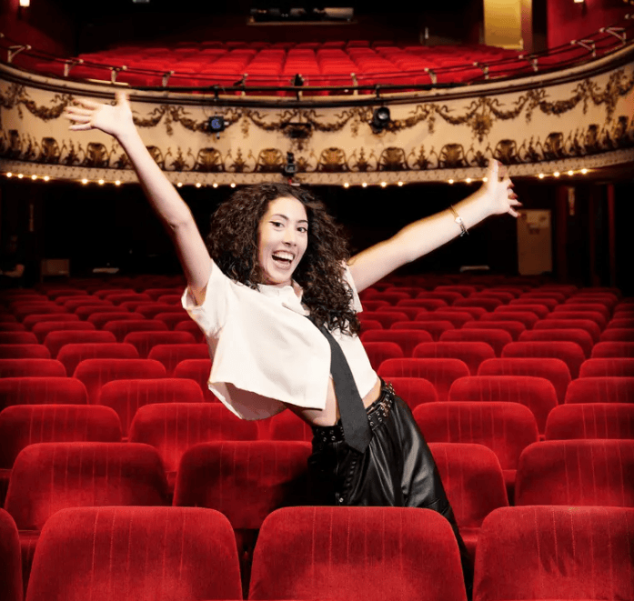 Jeune femme enthousiaste posant avec joie dans une salle de théâtre vide, entourée de sièges rouges et d’un décor intérieur élégant.