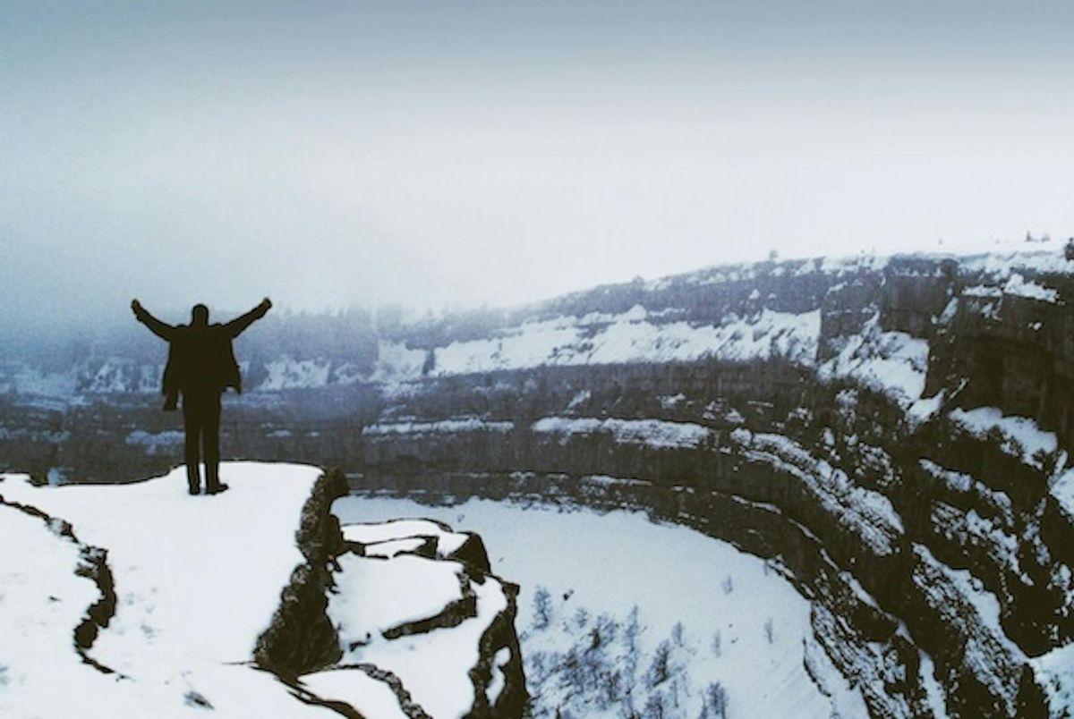 Un homme debout sur une falaise enneigée avec les bras levés, face à un paysage de canyon sous un ciel nuageux.