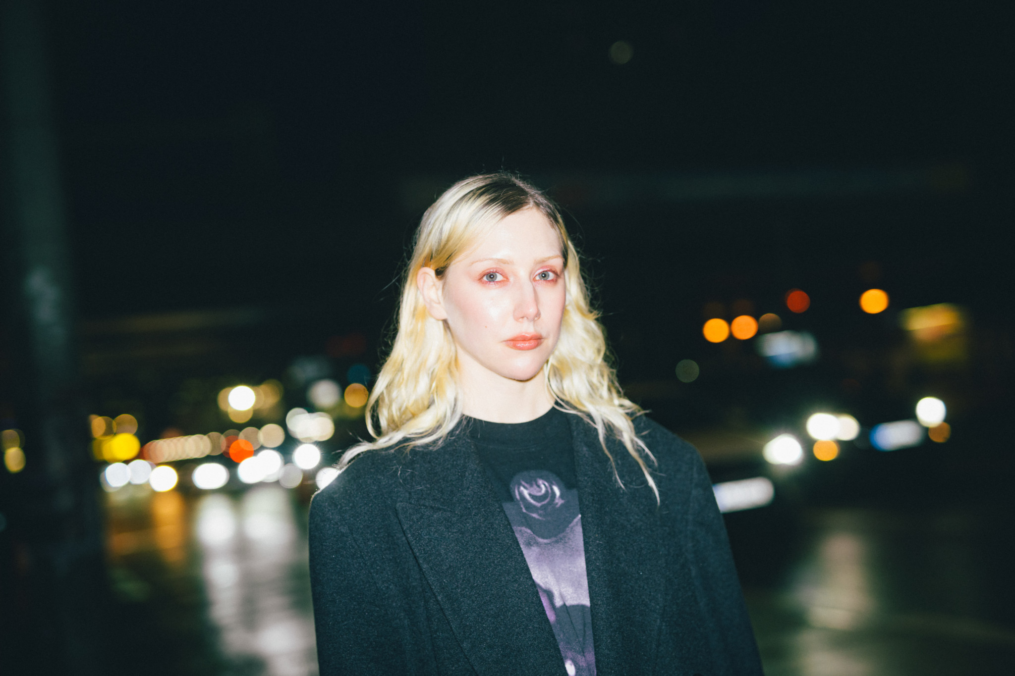 Ilona Hartmann mit blonden Haaren und schwarzem Jackett, nachts im Freien, verschwommene Stadtlichter im Hintergrund.