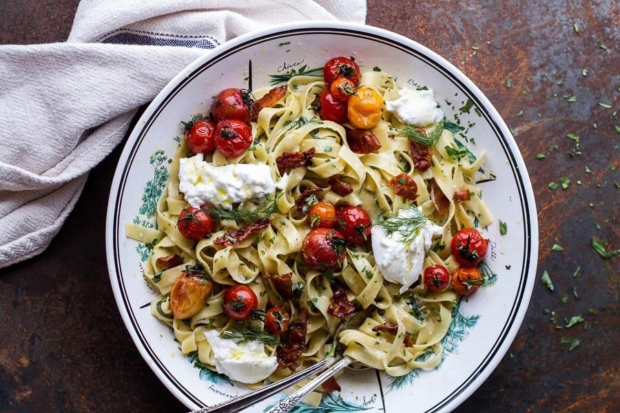 Teller mit Tagliatelle, gerösteten Kirschtomaten, Burrata und frischen Kräutern auf dunklem Untergrund.