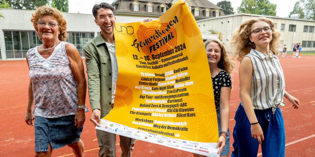 Vier freiwillige Helferinnen und Helfer halten auf einer Veranstaltung im Freien ein Banner für das Generationenfestival. Sie laufen auf einem Sportplatz der Kamera entgegen.