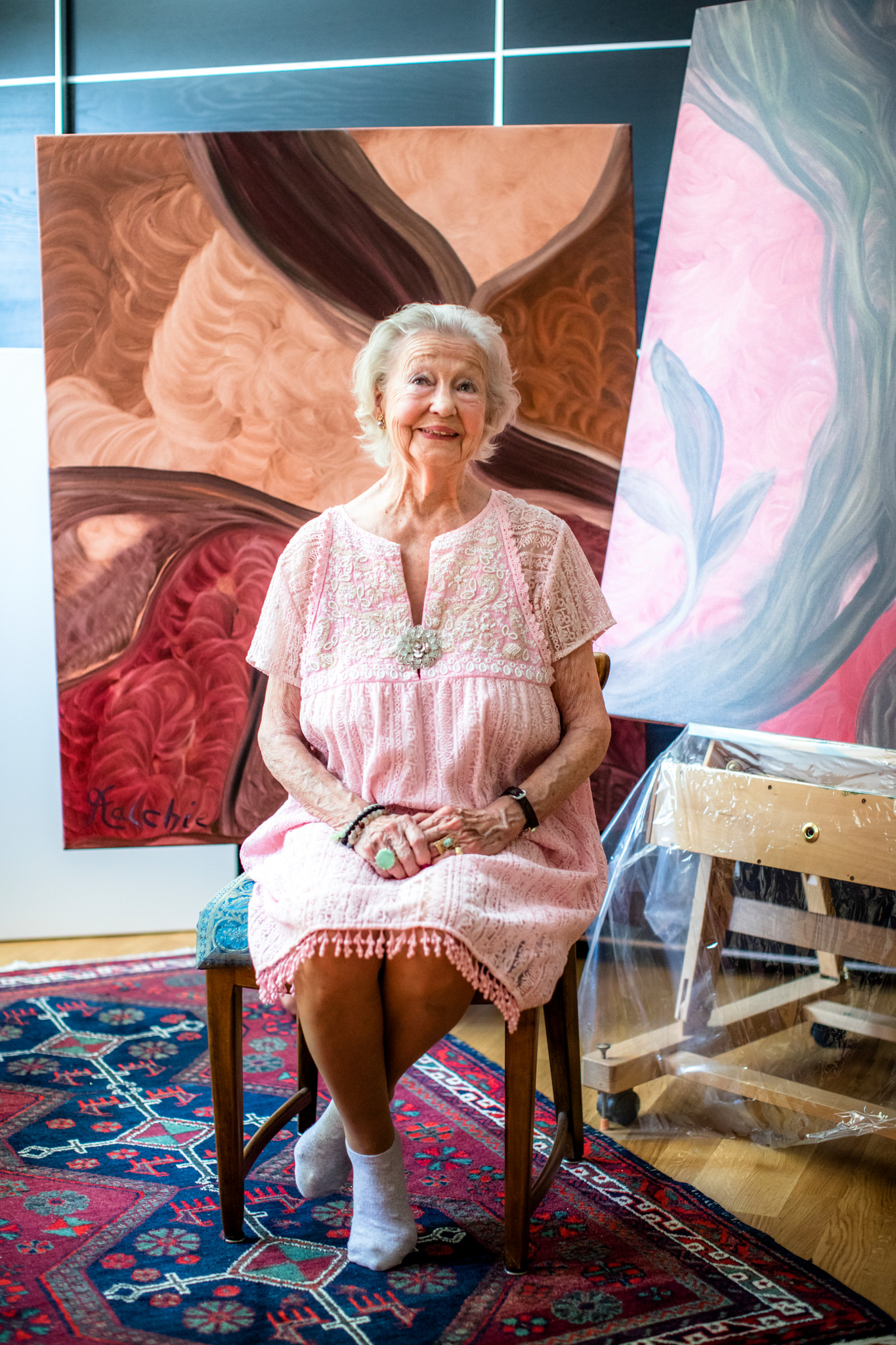 86-jährige Künstlerin in ihrer Wohnung sitzt vor zwei grossen Gemälden. Sie trägt ein rosa Kleid und sitzt barfuss auf einem Stuhl. Ein bunter Teppich liegt auf dem Boden.