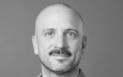 Homme chauve avec moustache portant une chemise en jean, souriant légèrement.