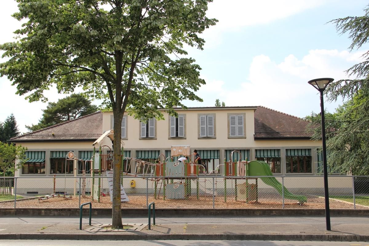 Cour d’école avec un bâtiment beige, abri à rayures vertes et aire de jeux clôturée sous un grand arbre.