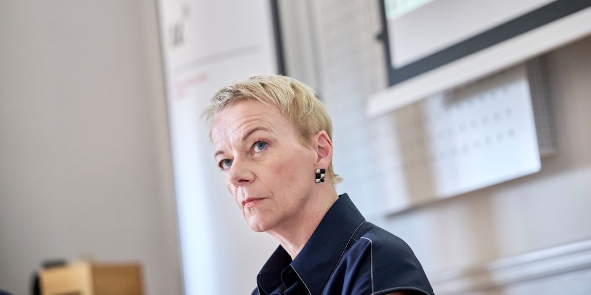 Frau mit kurzem blondem Haar sitzt in einem Büro und schaut ernst. Im Hintergrund sind ein Whiteboard und ein Projektor zu sehen.