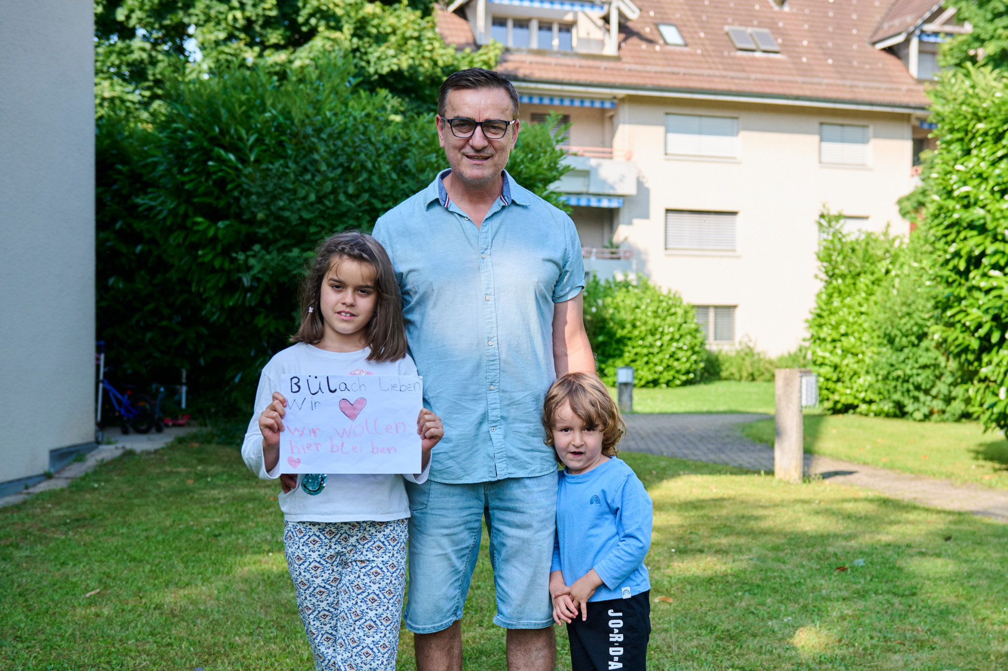 Ein Mann steht mit zwei Kindern in einem Garten. Das grössere Kind hält ein Schild mit der Aufschrift ’Bülach Leben ist wichtiger als der Gewinn.’