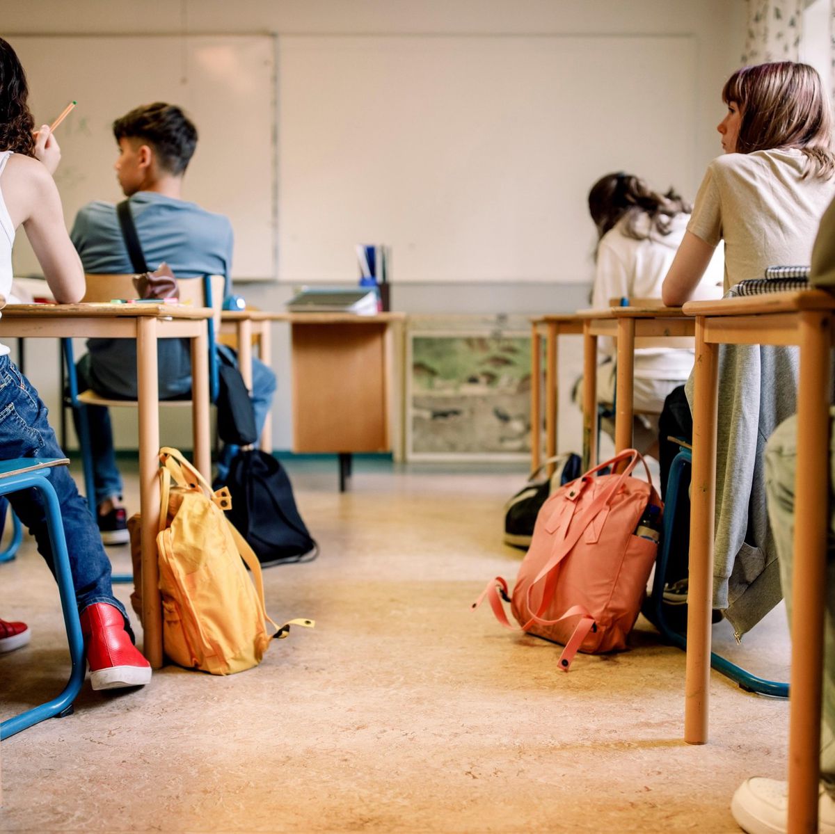 Salle de classe avec des élèves assis à leurs bureaux, sacs à dos colorés au sol.