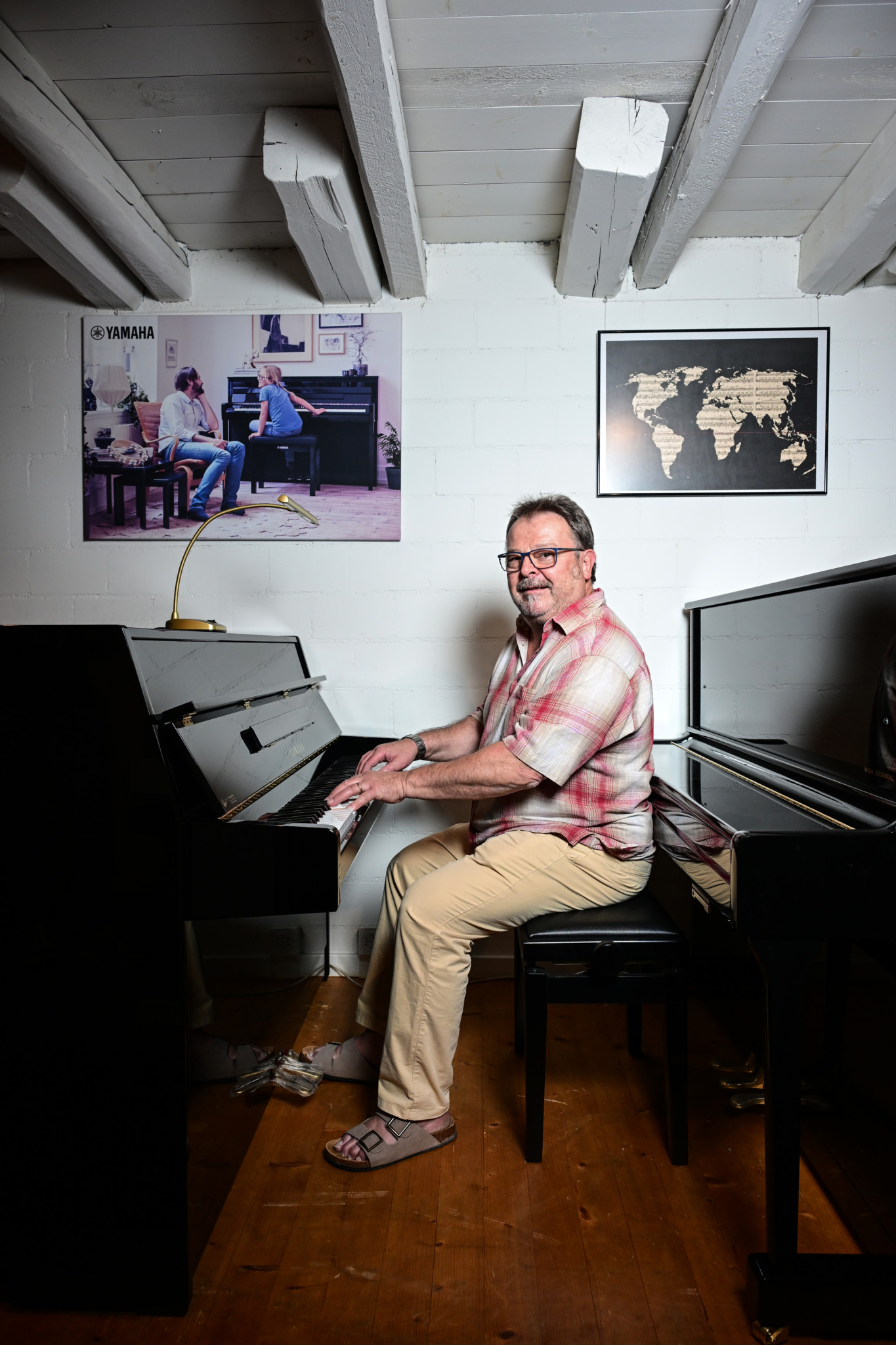 Roland Schmidt sitzt lächelnd an einem schwarzen Klavier in seinem Musighuus in Wiesendangen, im Hintergrund ein Yamaha-Poster und eine Weltkarte.