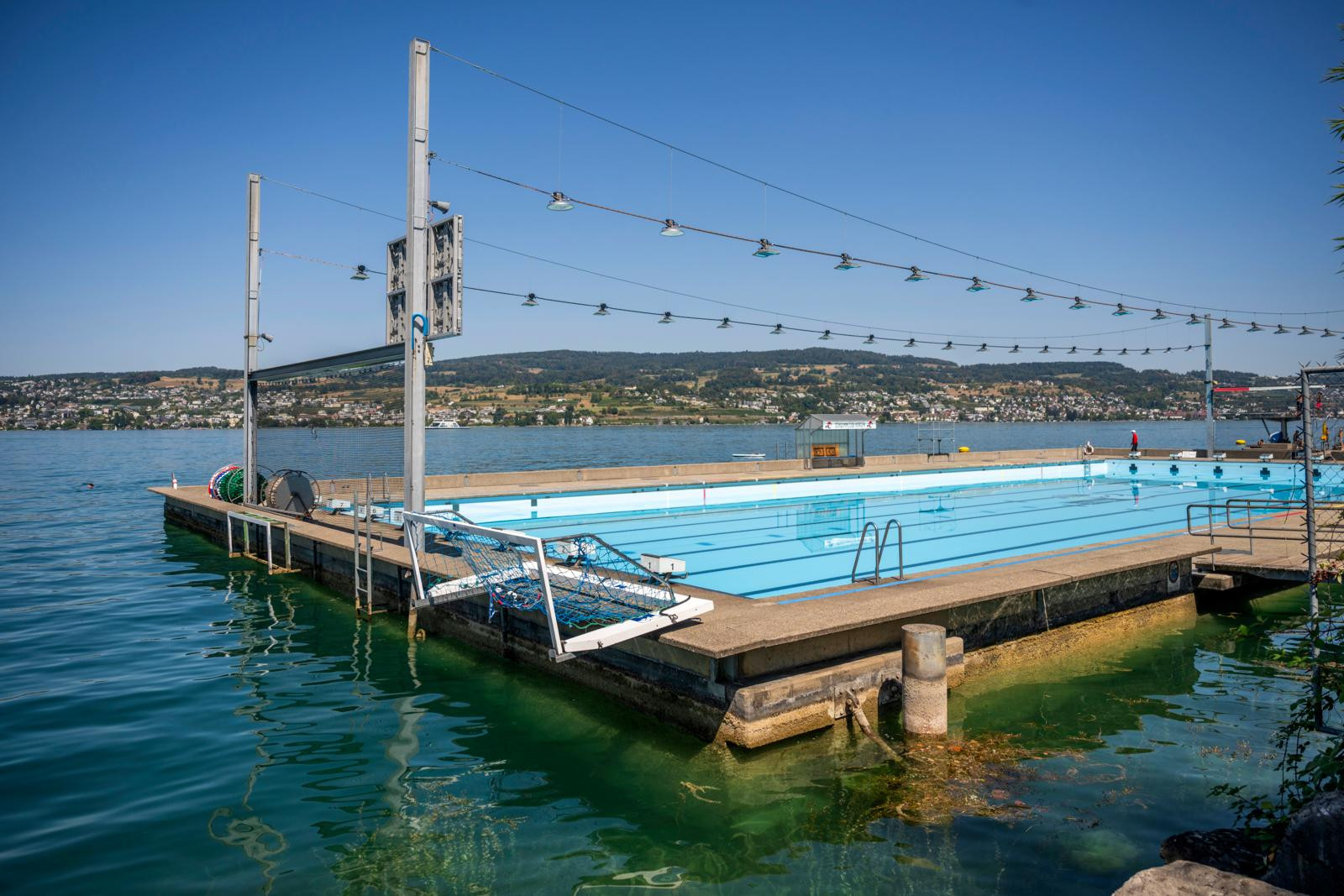 Schwimmbad auf einem Steg direkt am See, mit türkisblauem Wasser, Lichterketten und Hügeln im Hintergrund bei strahlend blauem Himmel.
