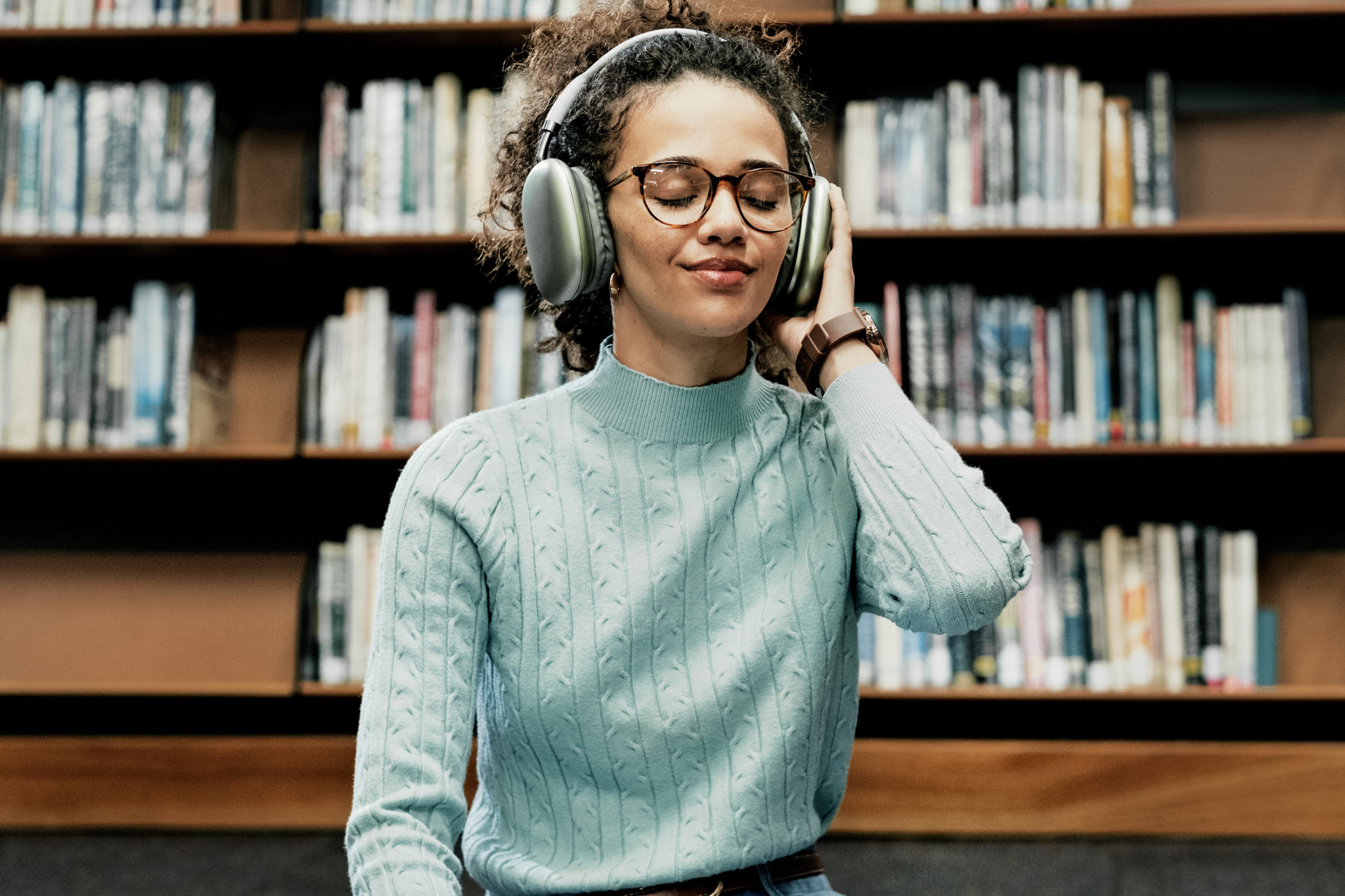 Lächelnde Frau mit Kopfhörern und Brille hört Hörbuch in einer Bibliothek, umgeben von Bücherregalen.