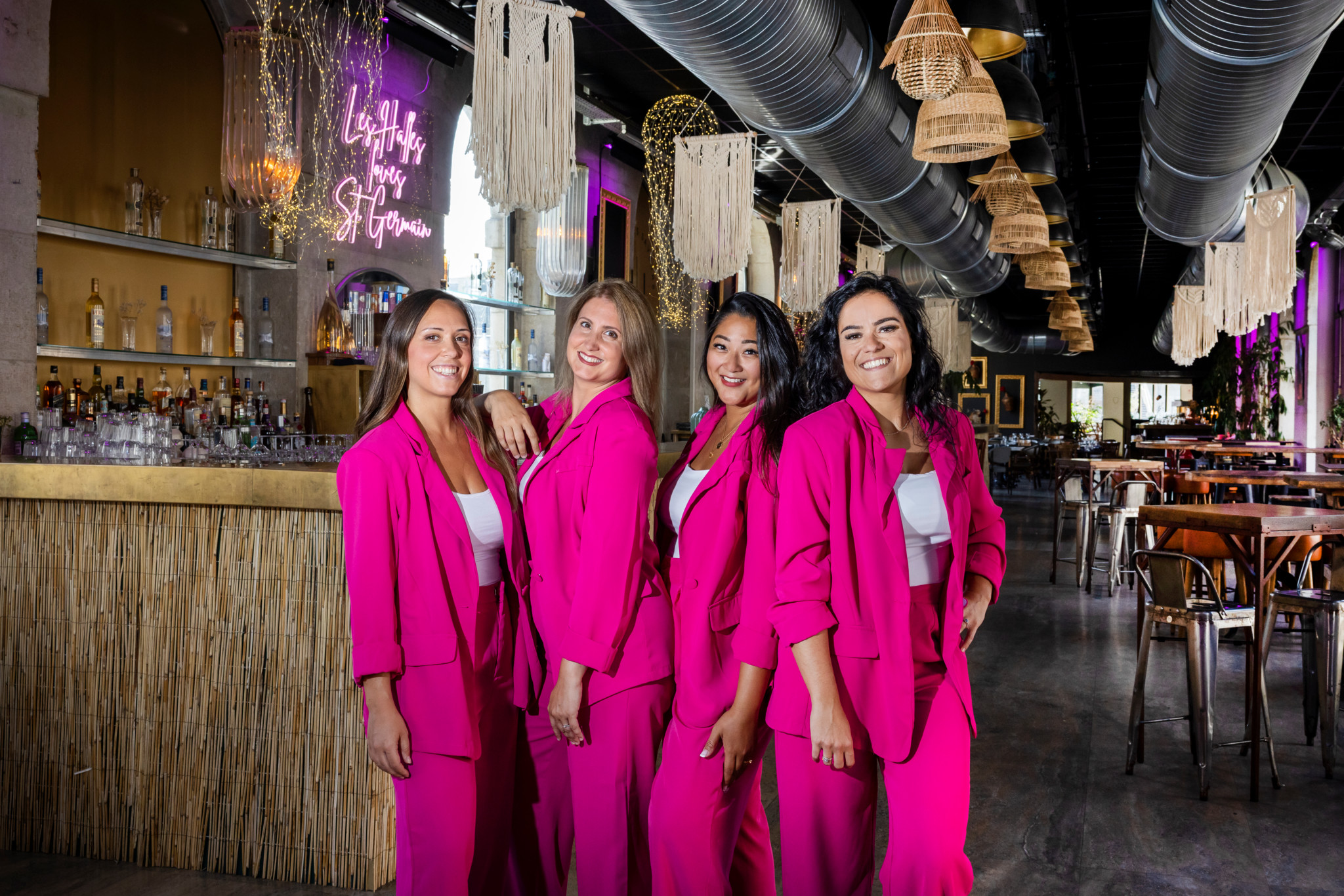 Deborah, Laetitia, Rachelle et Dorothee, fondatrices de ’Give Mama a Drink’, posent ensemble dans un bar, vêtues de costumes roses vifs.