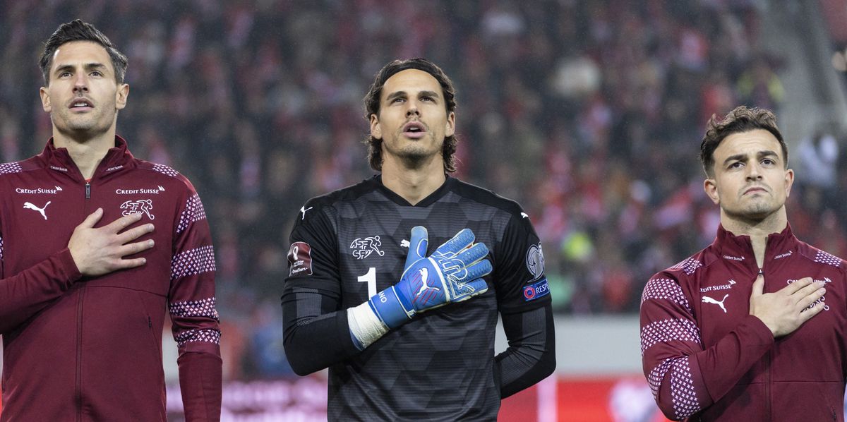 Trois joueurs de football suisses chantent l'hymne national avant un match, main sur le cœur. Le joueur central porte un maillot de gardien noir, tandis que les joueurs à sa gauche et à sa droite portent des maillots de terrain rouges avec des motifs blancs.