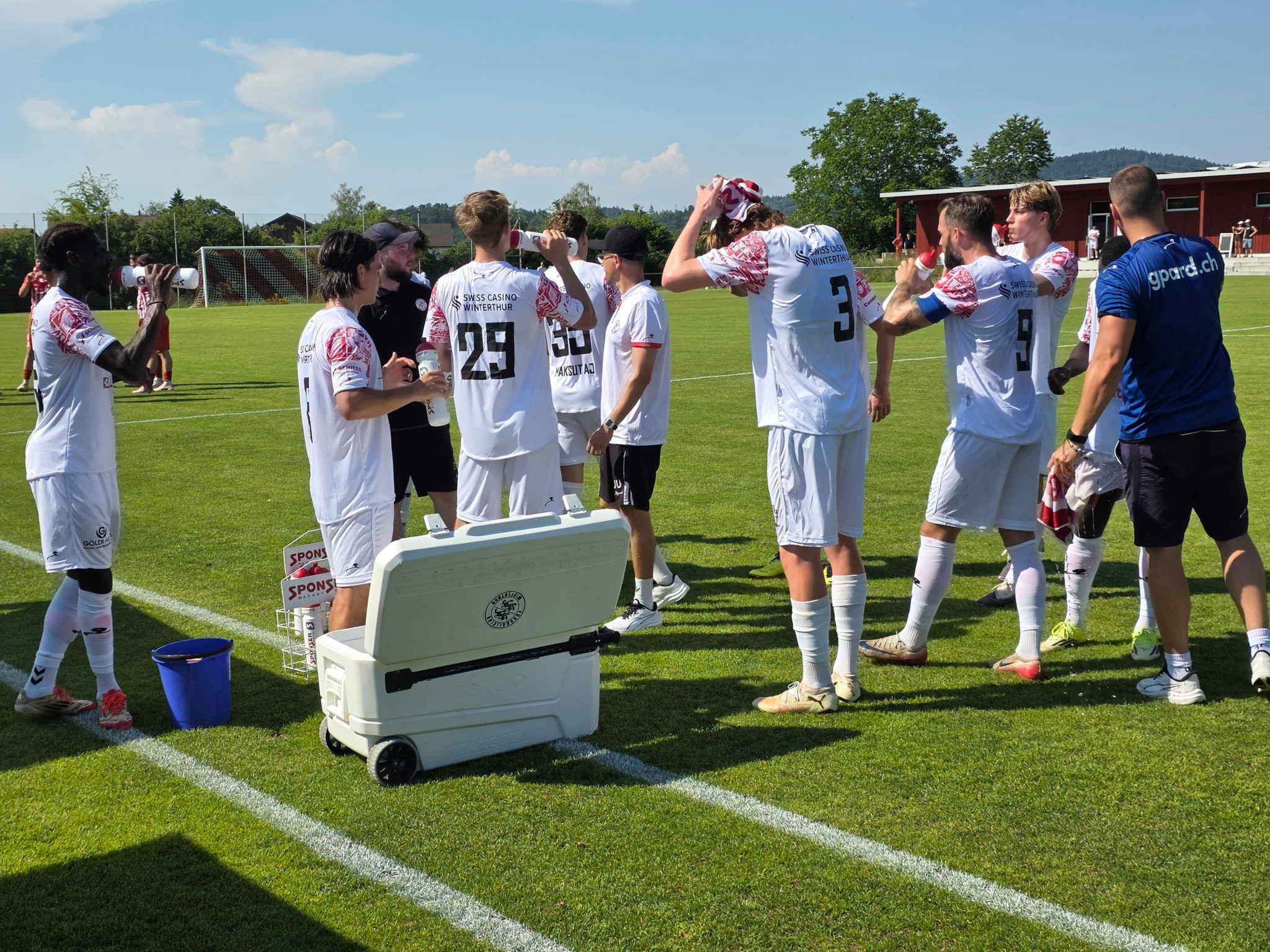 Fussballspieler in weissen Trikots machen eine Pause am Spielfeldrand, neben einer Kühlbox, bei sonnigem Wetter.