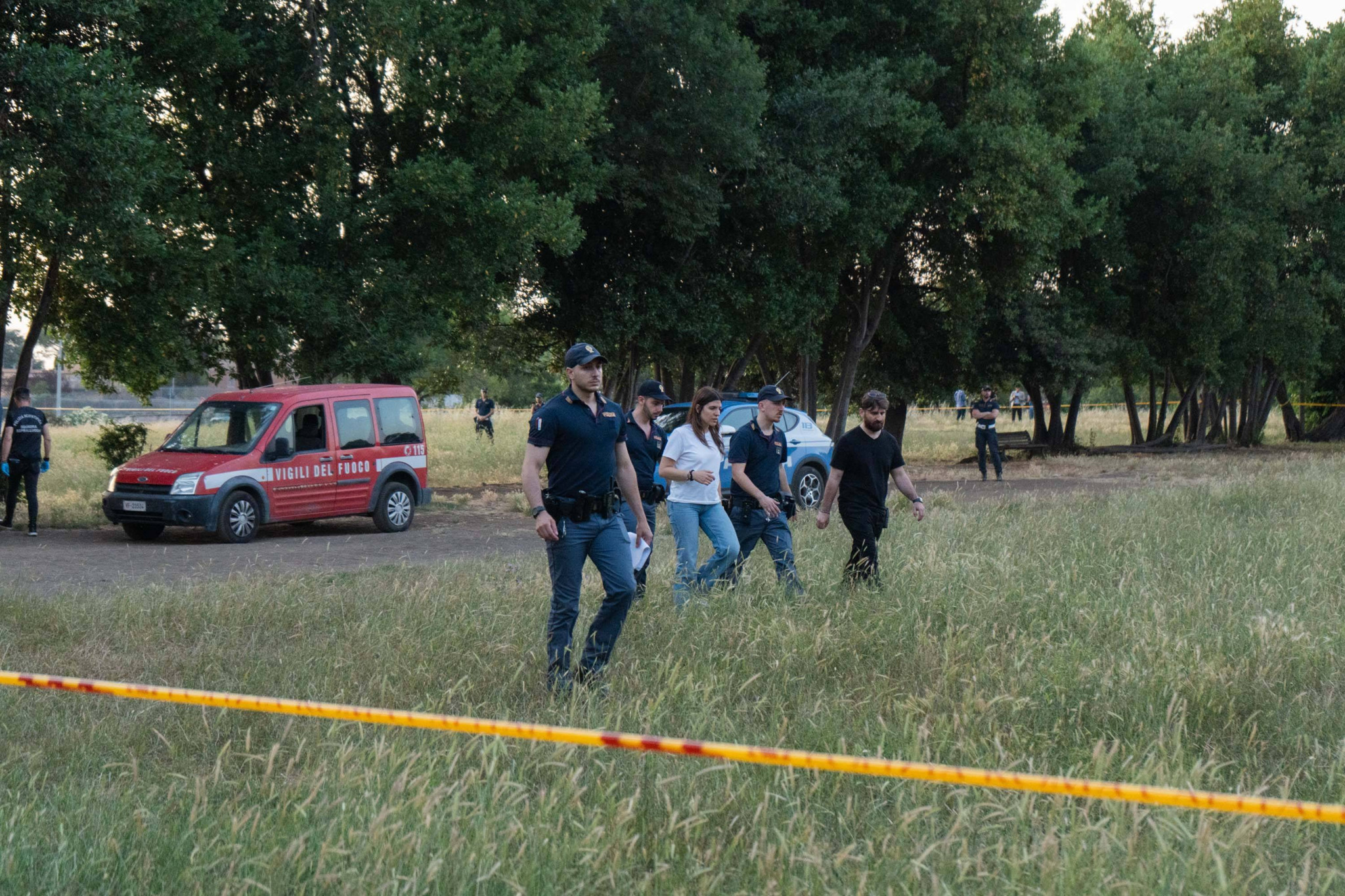 Polizeibeamte durchsuchen ein Feld in Villa Doria Pamphilj, mit einem Feuerwehrfahrzeug im Hintergrund. Ein Vorfall mit einer verstorbenen Frau und einem Neugeborenen wird untersucht.