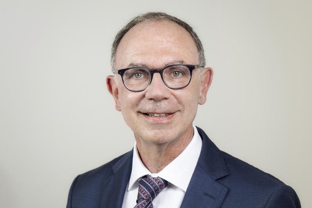 Portrait d’une personne en costume et cravate, portant des lunettes, souriant légèrement devant un fond neutre.