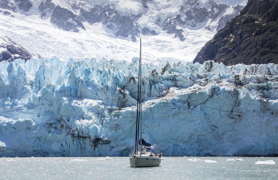 Un voilier navigue devant un glacier majestueux entouré de montagnes enneigées.