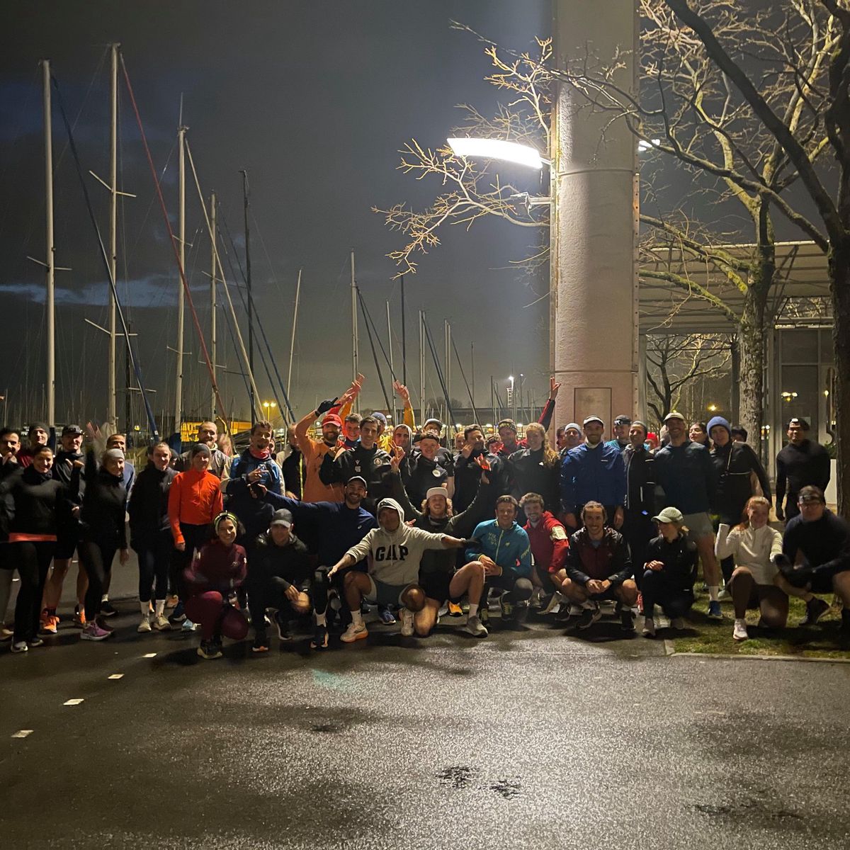 Groupe de personnes posant devant un port de plaisance la nuit, sous des lampadaires avec des voiliers à l’arrière-plan.