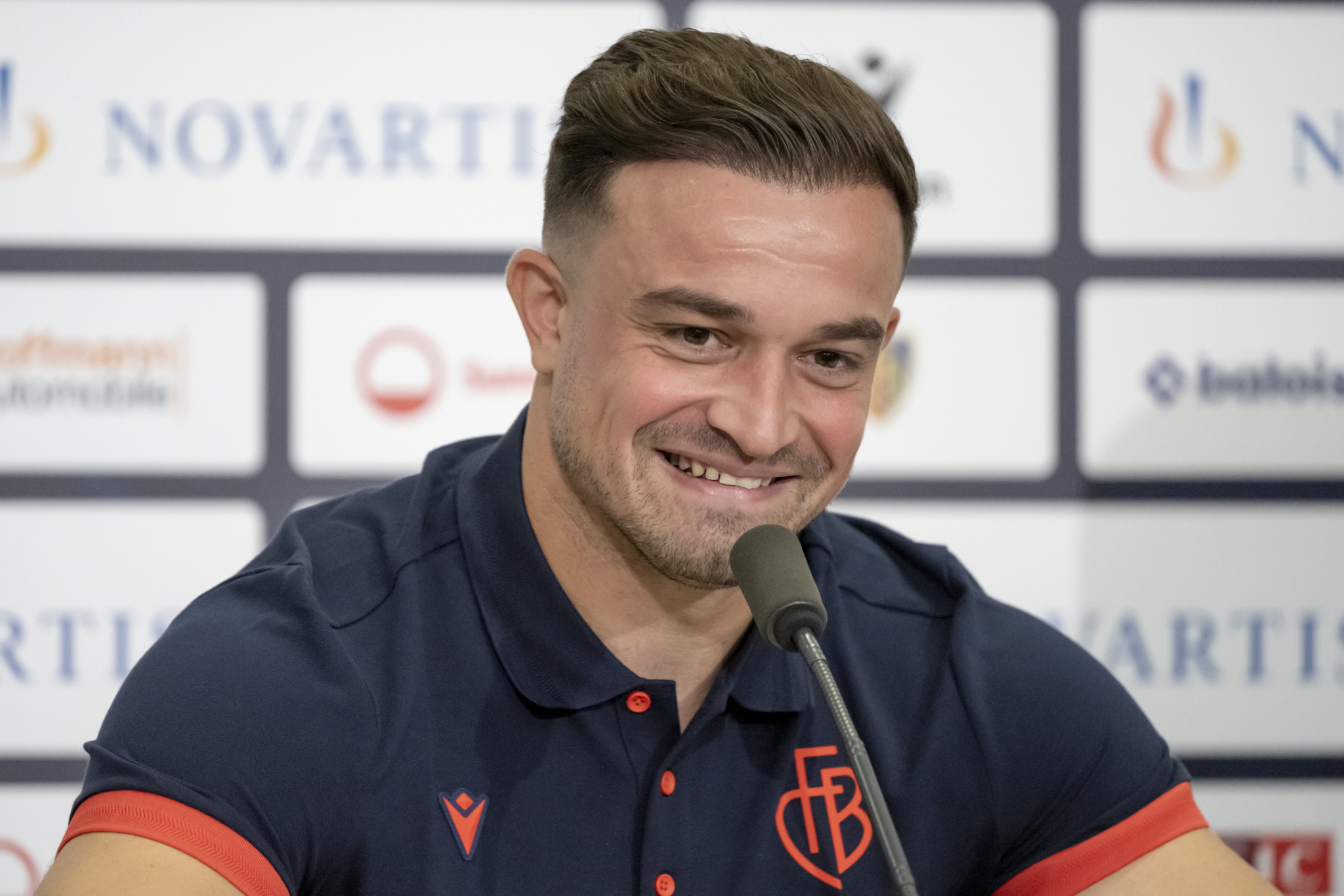 Xherdan Shaqiri spricht lächelnd auf einer Medienkonferenz in Basel, mit der Logo-Wand von FC Basel und Sponsoren im Hintergrund.