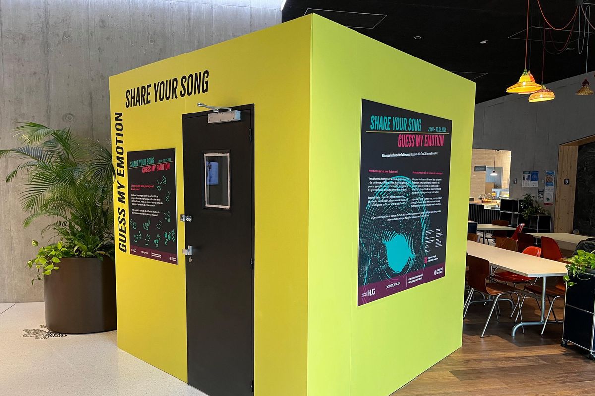 Cabine jaune intitulée ’Share Your Song’ dans un espace intérieur avec tables, chaises et plantes.